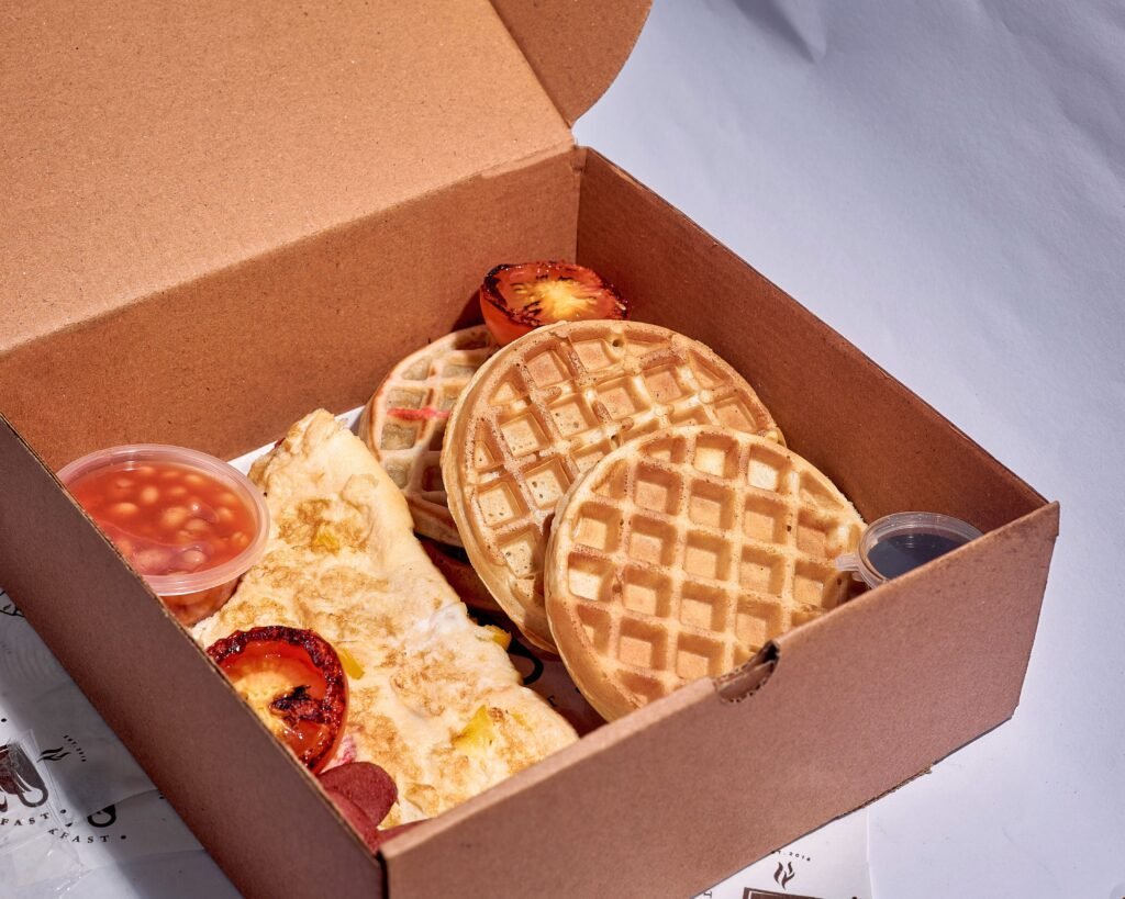 Cardboard takeout box featuring waffles, omelette, and grilled tomatoes, perfect breakfast meal.